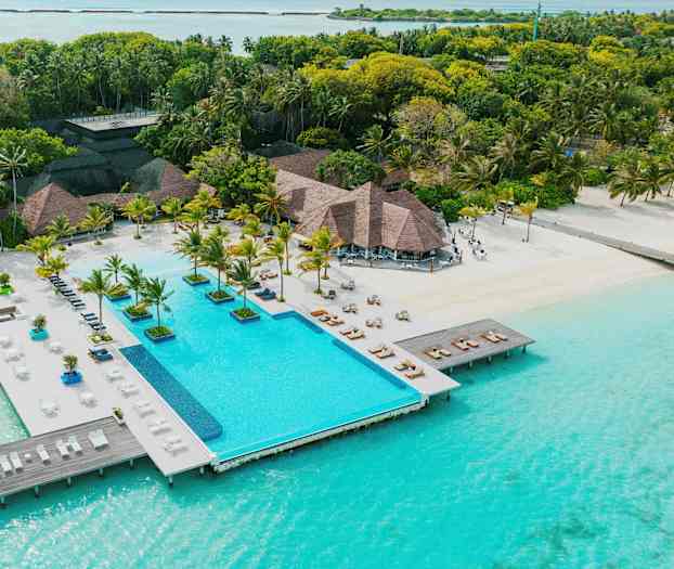 Et resort med en pool, der ligger lige ved turkisblåt vand, omgivet af palmer og grønt. (AI-genereret tekst)