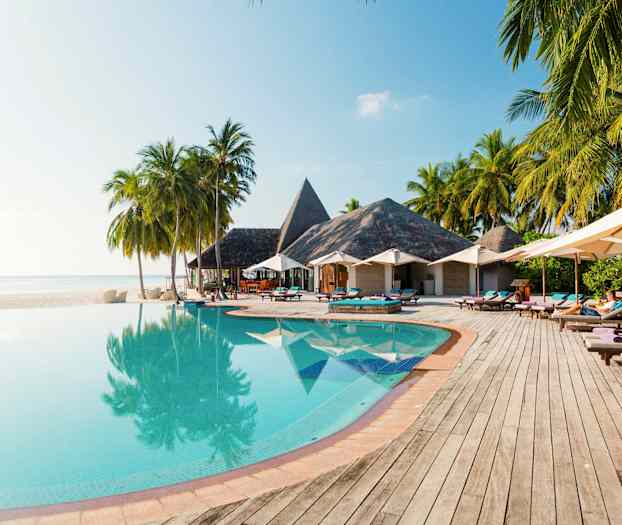 En infinitypool med træer og parasoller ved siden af, samt bygninger med stråtag i baggrunden. (AI-genereret tekst)