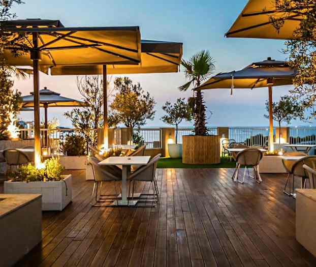 Billede af en udendørs terrasse med trægulv, borde og stole under parasoller, omgivet af grønne omgivelser i skumringslys. (AI-genereret tekst)