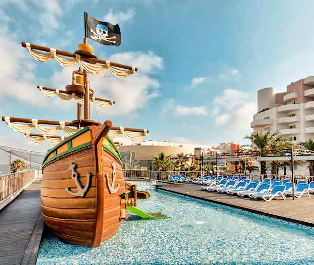 Billede af poolområdet med et lege-piratskib og liggestole i baggrunden. (AI-genereret tekst)