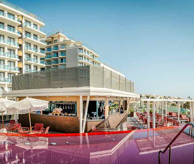 Et hotel med altaner og en pool foran, samt en bar og siddepladser under parasoller. (AI-genereret tekst)