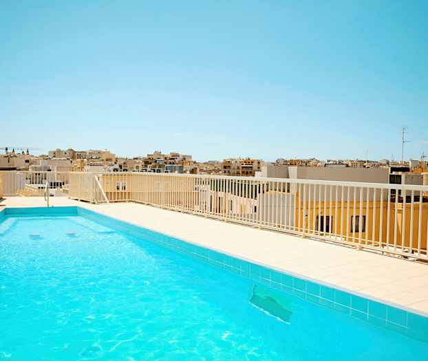 En udendørs pool med klart vand på en tagterrasse. I baggrunden ses bybebyggelse under en klar blå himmel. (AI-genereret tekst)