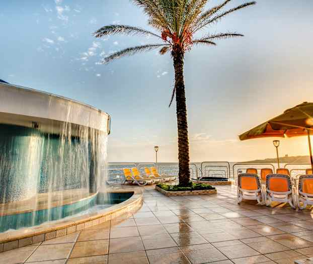 En pool med et vandfald ved siden af en palme, liggestole og udsigt over havet på en solrig dag. (AI-genereret tekst)