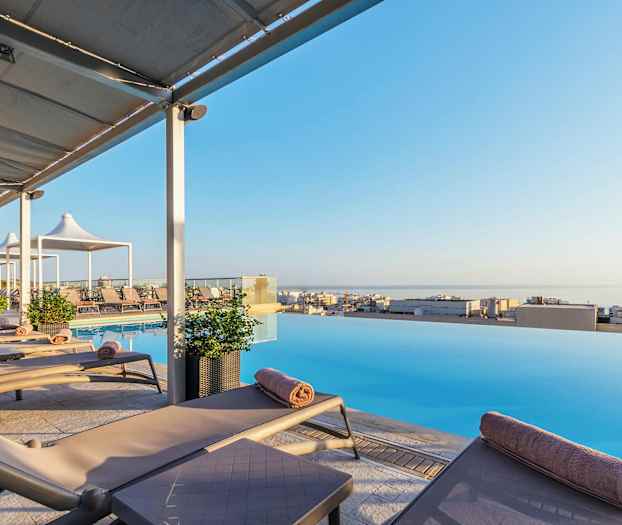 Liggestole ved en infinity-pool med udsigt over en by og havet. (AI-genereret tekst)