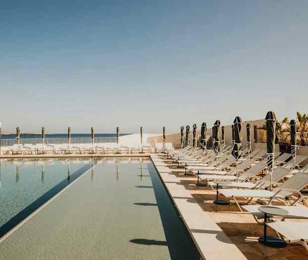 Billede af en swimmingpool med liggestole og parasoller i et solrigt udendørsområde. (AI-genereret tekst)