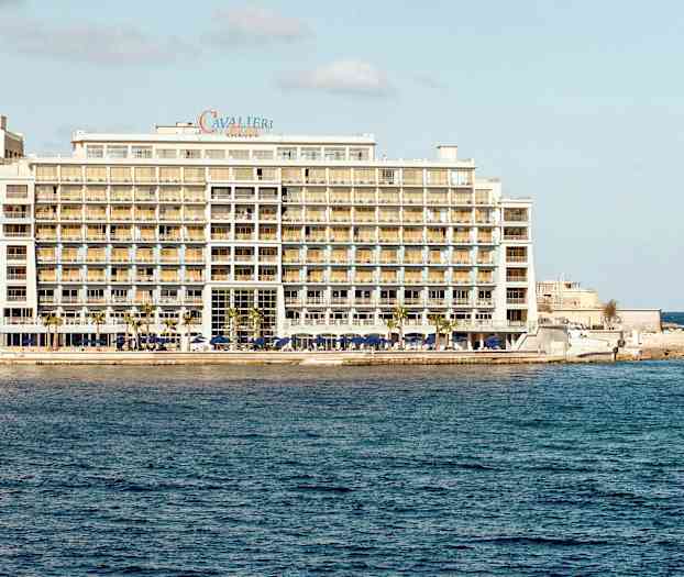 Et stort hotel ved havet med flere etager og altaner samt en lille båd i vandet tæt på hotellet. (AI-genereret tekst)