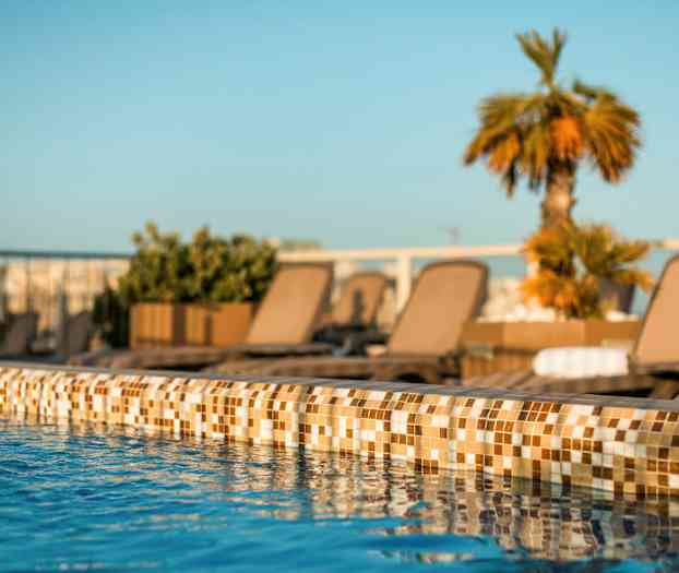 Nærbillede af en poolkant med klart blåt vand og liggestole samt planter i baggrunden. (AI-genereret tekst)