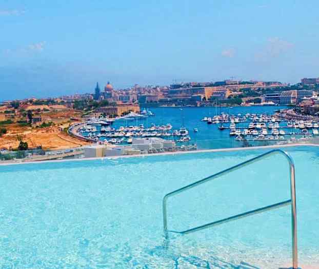 Udsigt over en pool med rækværk, hvor en havn med mange både og bymiljø ses i baggrunden. (AI-genereret tekst)