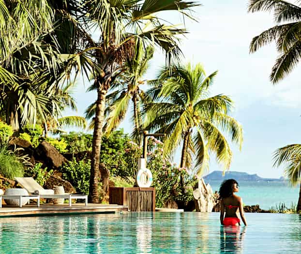 En person sidder i en pool med udsigt til tropiske palmer og havlandskab. (AI-genereret tekst)