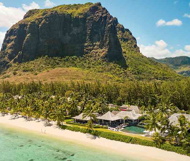 Et stort bjerg ses i baggrunden af en strand med turkis vand i forgrunden. På stranden står palmer og bygninger med stråtag. (AI-genereret tekst)