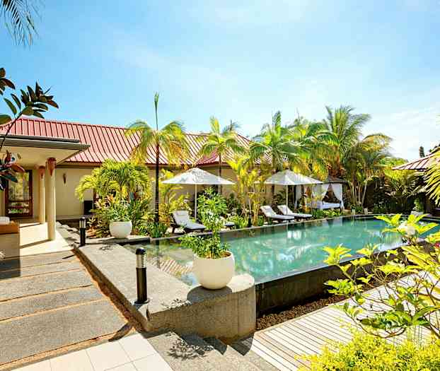 Et åbent miljø med en pool omgivet af liggestole og parasoller, med grøn beplantning og bygninger i baggrunden. (AI-genereret tekst)