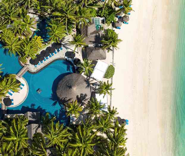 Billedet viser et luftperspektiv over en strand med hvidt sand, turkis vand og en pool omgivet af palmer. (AI-genereret tekst)