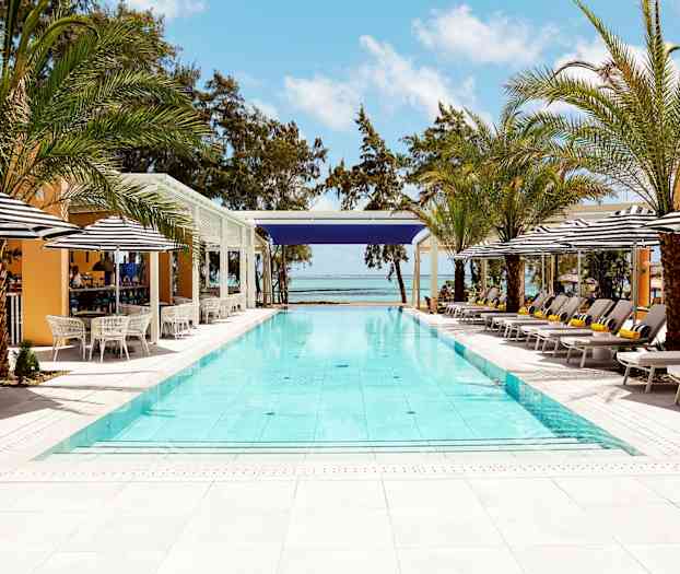 Udendørs pool omgivet af liggestole, parasoller og palmer med hav og blå himmel i baggrunden. (AI-genereret tekst)