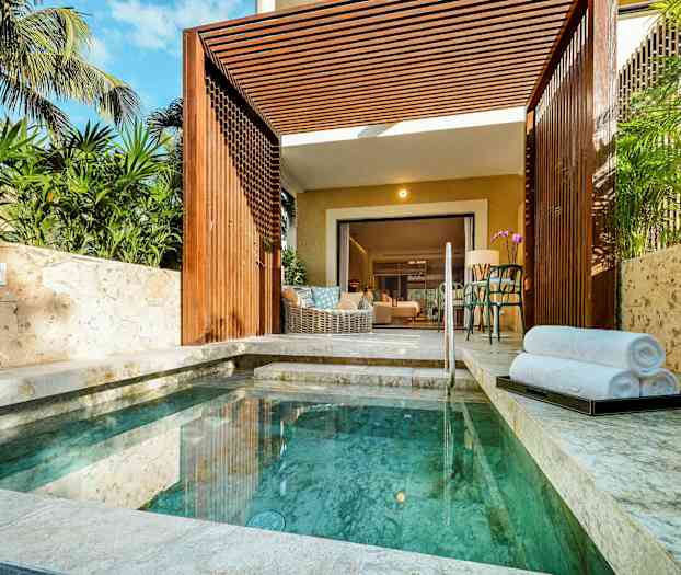 En lille pool omgivet af stenbelægning og grønne planter med en åben terrasse, der fører ind til et værelse. (AI-genereret tekst)