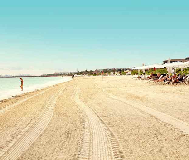 En sandstrand med parasoller og liggestole, en person står ved vandet. (AI-genereret tekst)