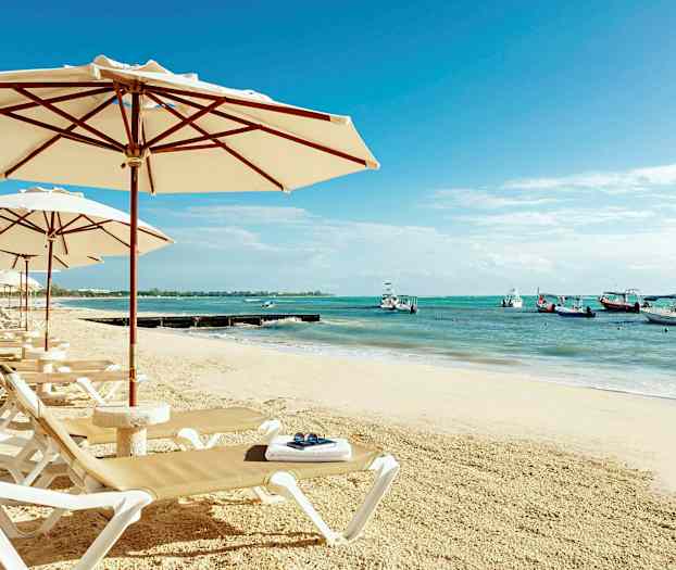 En sandstrand med solsenge og parasoller. I baggrunden ses havet med både. (AI-genereret tekst)