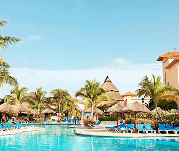 Billedet viser en udendørspool omgivet af liggestole med blå puder og parasoller. I baggrunden ses palmer og bygninger med orange tage. (AI-genereret tekst)