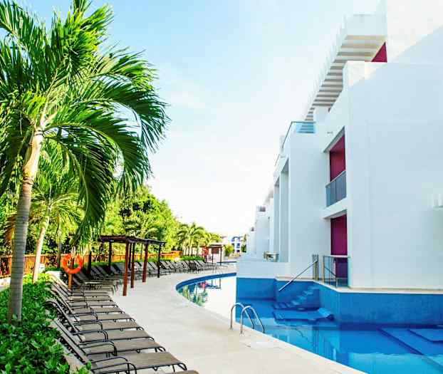 Billede af en swimmingpool omgivet af liggestole, palmer og moderne hvide bygninger. (AI-genereret tekst)