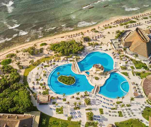 Luftfoto af et feriested ved havet med flere swimmingpools, omgivet af grønne områder og bygninger med stråtag. (AI-genereret tekst)