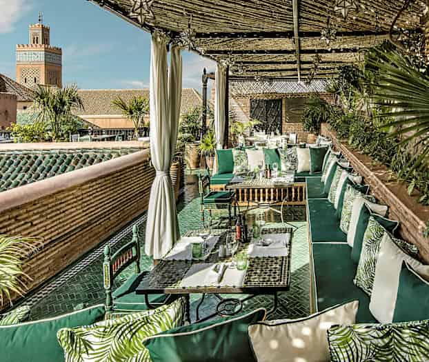 En udendørs terrasse med grønne siddepladser og planter, omgivet af tag og murstensvægge. En minaret er synlig i baggrunden. (AI-genereret tekst)
