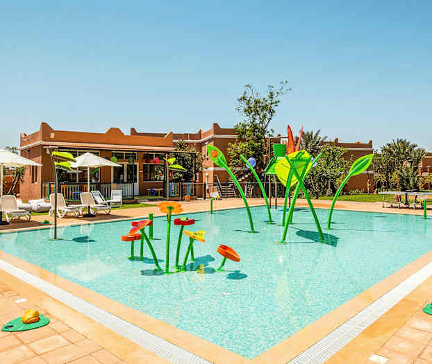 Udendørs swimmingpool med vandlegeudstyr formet som blomsterlignende figurer i grøn og orange, omgivet af solvogne og parasoller. (AI-genereret tekst)