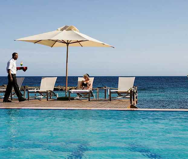 En pool ved havet med liggestole og parasol samt en person, der serverer en drink. (AI-genereret tekst)