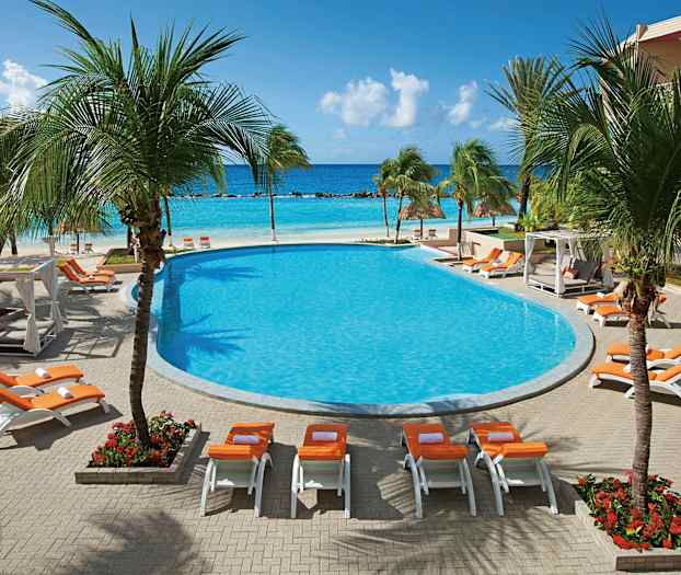 Et poolområde med liggestole og parasoller, omgivet af palmer, med udsigt over havet i baggrunden. (AI-genereret tekst)