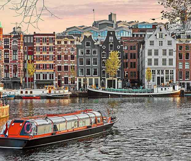 Flere bygninger med traditionelle facader ved en kanal, hvor en sightseeingbåd passerer i forgrunden. (AI-genereret tekst)