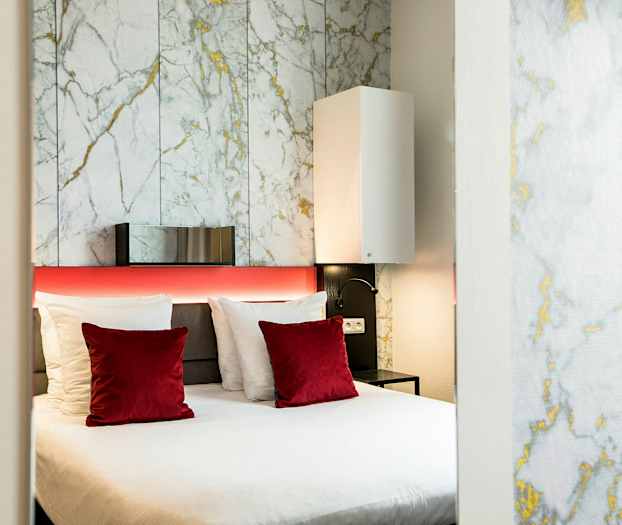 Et moderne hotelværelse med en dobbeltseng, hvide og røde puder samt en bagvæg med marmormønster. (AI-genereret tekst)