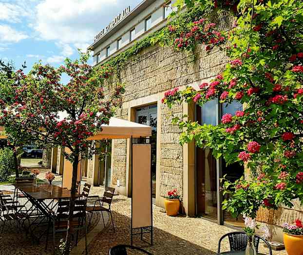 En udendørsterrasse med blomster og borde ved siden af en stenbygning. (AI-genereret tekst)