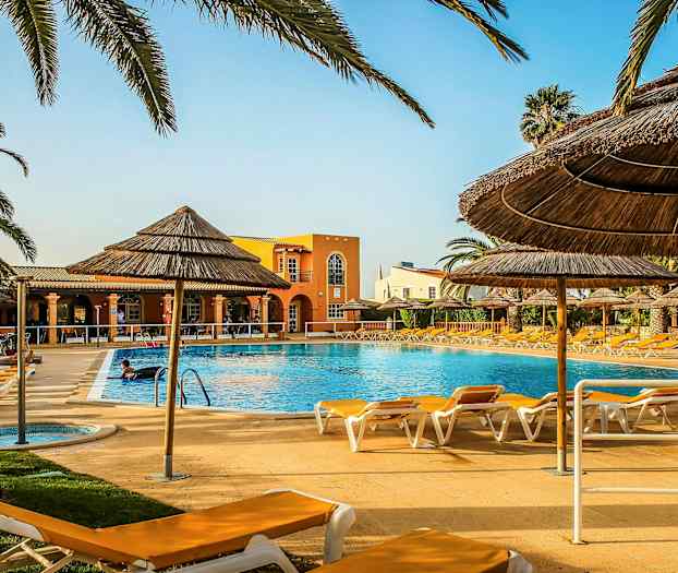 Udendørs swimmingpool omgivet af liggestole og halmparasoller med en bygning i baggrunden. (AI-genereret tekst)