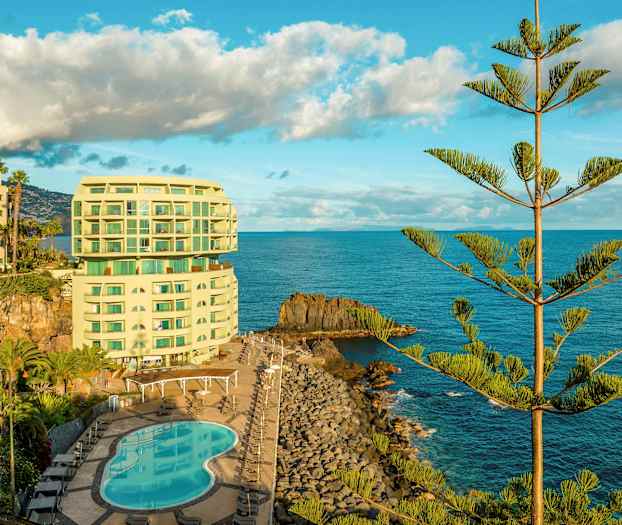 Et hotel ved kysten med en pool og omgivet af hav og træer. (AI-genereret tekst)