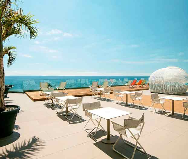 En udendørs terrasse med hvide stole og borde, skyggefulde palmer, udsigt over havet samt en hvid kuppelformet struktur i baggrunden. (AI-genereret tekst)