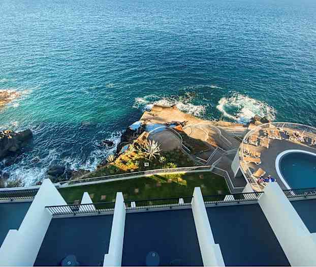 Luftfoto af et anlæg ved kysten med blåt hav, klipper, pool og solsengsområde. (AI-genereret tekst)