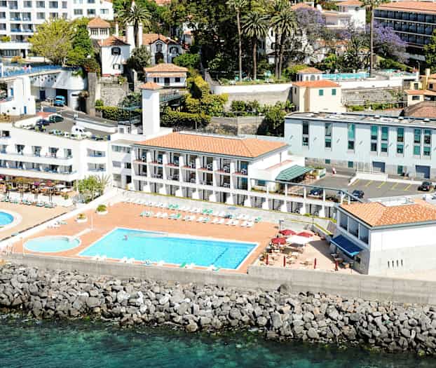 Luftfoto af et hotel med pools, liggestole og bygninger tæt på havet. (AI-genereret tekst)