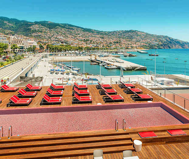 Poolområde med liggestole og udsigt over havn og by. (AI-genereret tekst)