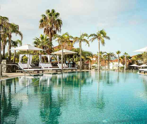 En luksuriøs pool med liggestole og parasoller, omgivet af palmer og bygninger i baggrunden. (AI-genereret tekst)