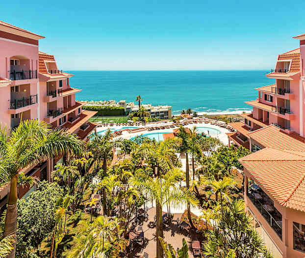 Udsigt over hotelområde med lyserøde bygninger, palmer og en pool med liggestole nær havet. (AI-genereret tekst)