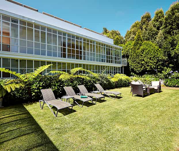 Billede af en have med liggestole, siddegruppe og grønne planter foran en bygning med store vinduer. (AI-genereret tekst)