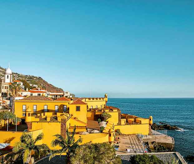 Gul bygning ved havet med omkringliggende grønne områder og bakgrunden med bakker. (AI-genereret tekst)