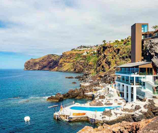 Kystlandskab med en moderne bygning og poolområde ved klipperne, omgivet af havet og frodige skråninger. (AI-genereret tekst)