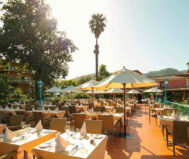 Udendørs serveringsområde med dækkede borde, parasoller og en frodig omgivelse. (AI-genereret tekst)