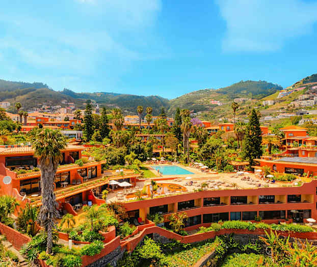 Et anlæg med terrakottafarvede bygninger, swimmingpool og frodig beplantning, beliggende i en dal omgivet af bakker. (AI-genereret tekst)