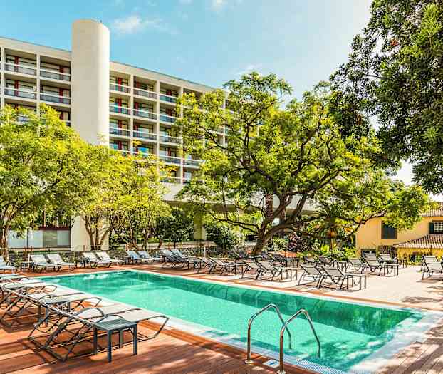 Udendørs pool med liggestole omgivet af træer og bygninger. (AI-genereret tekst)