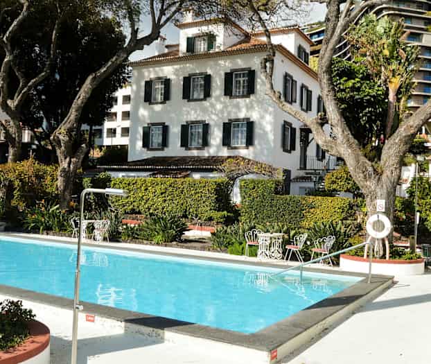 Billede af en udendørs pool med vand og omkringliggende træer og buske, med hvide bygninger i baggrunden. (AI-genereret tekst)