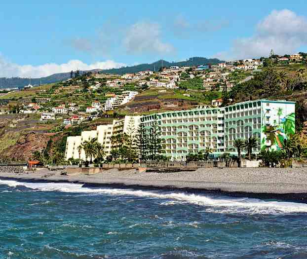 Hotelbygninger ved en strand med havebølger og en baggrund af grønne bakker og boliger. (AI-genereret tekst)
