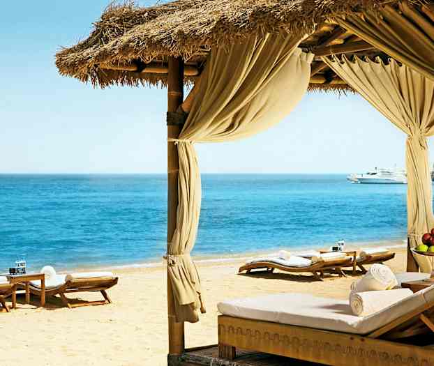 En strand med liggestole og en pavillon vender ud mod et klart hav. (AI-genereret tekst)