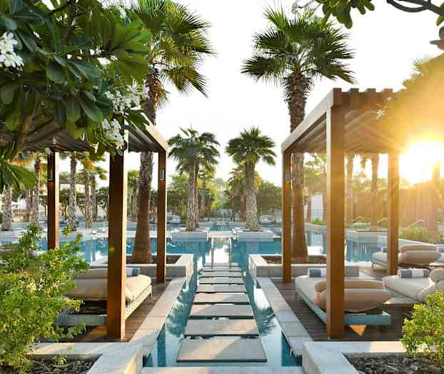 Et haveanlæg med pools, liggestole og palmer under pergolatag med solskin i baggrunden. (AI-genereret tekst)