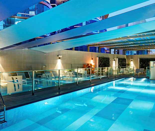 En indendørs pool med belysning. Rundt om poolen er der liggestole og glasværn. (AI-genereret tekst)