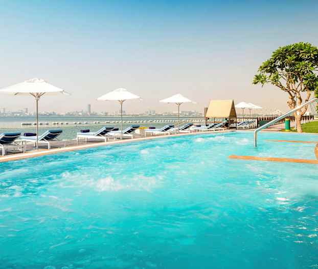 Udendørs pool med liggestole og parasoller ved havet, byens silhuet er synlig i baggrunden. (AI-genereret tekst)